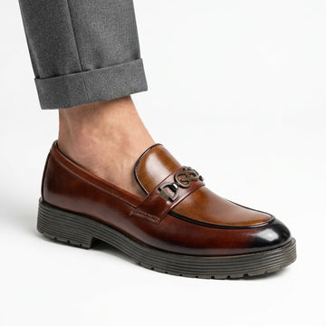 Tan burnished leather chunky-sole loafer with gunmetal logo hardware worn with grey trousers