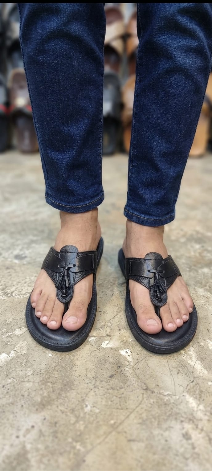 Black leather tassel medallion T-bar flip sandal pair standing upright against kraft gift box on rustic wooden table with folded dark denim jeans