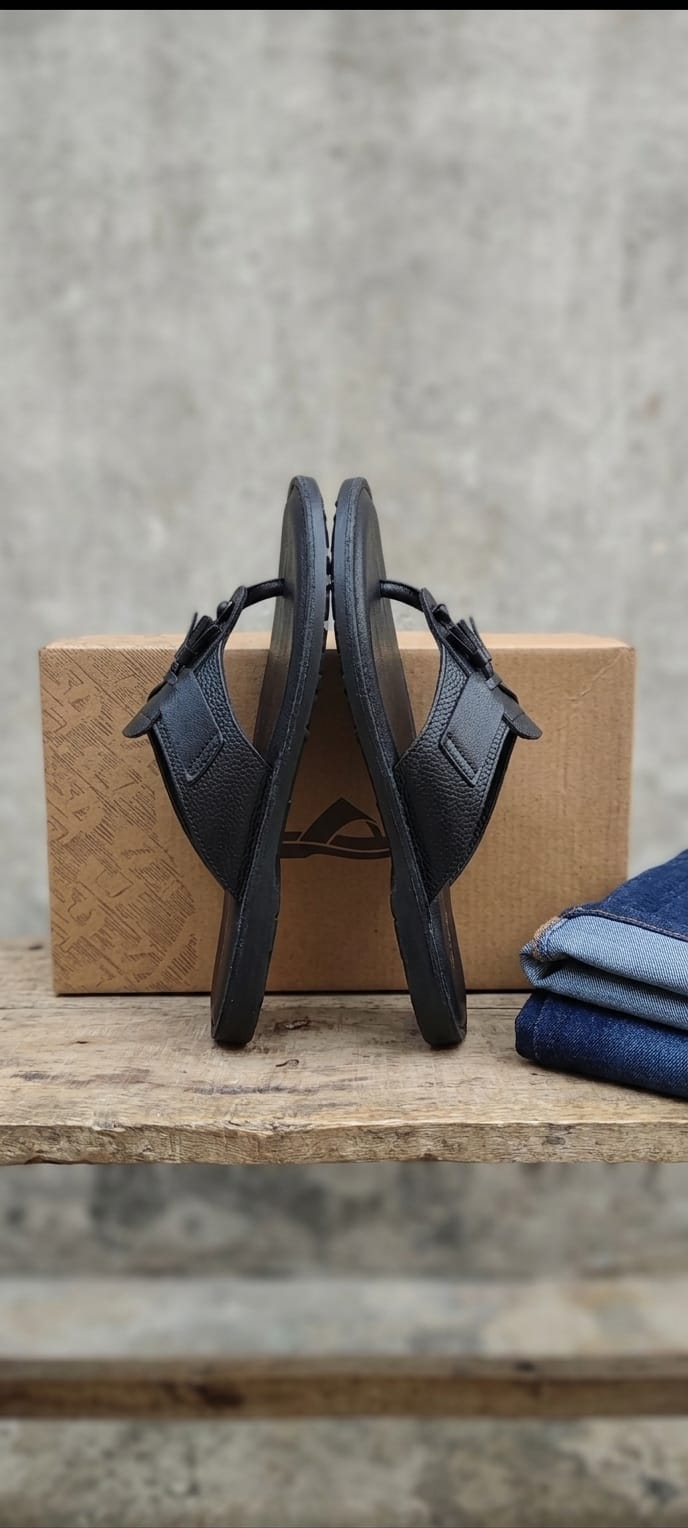 Black leather tassel medallion T-bar flip sandal pair standing upright against kraft gift box on rustic wooden table with folded dark denim jeans