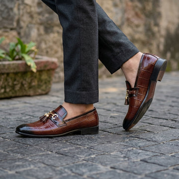Brown croc-embossed tassel horsebit loafer worn on cobblestone street with dark charcoal trousers