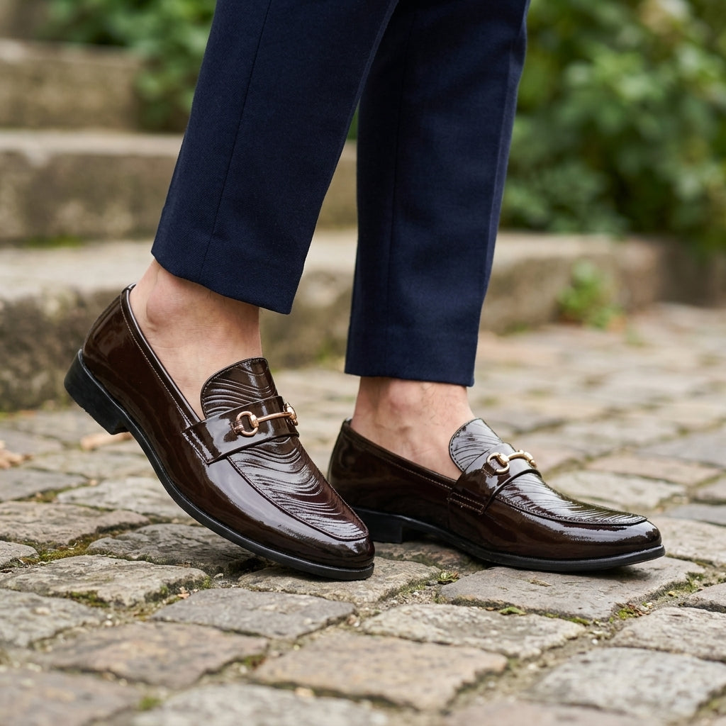 Dark brown patent wave-texture horsebit loafer worn outdoors on cobblestone with navy trousers