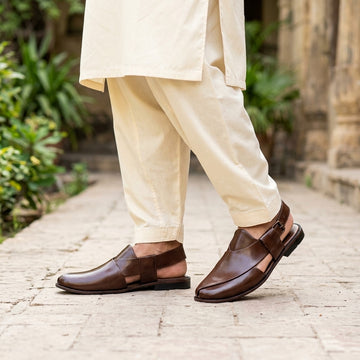 Brown leather plain-toe Peshawari slingback chappal worn on cobblestone pathway with cream shalwar kameez
