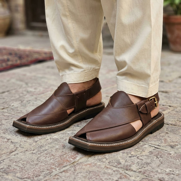 Dark brown cross-strap Kaptaan Peshawari chappal worn on stone courtyard with cream shalwar kameez gold buckle visible